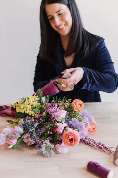 Florist smiling while wrapping a spring bouquet in a soft mauve ribbon, featuring bright orange ranunculus, yellow mimosa, purple statice, and blush sweet peas.