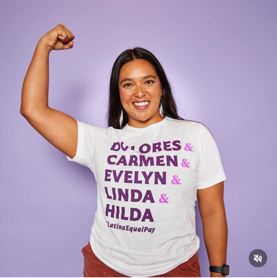 Member of LAU raising her fist and smiling confidently in a white T-shirt listing iconic Latina names, standing against a purple backdrop.