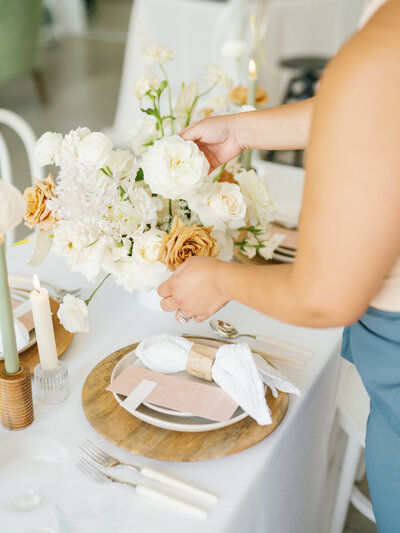 Eventive Designs wedding planner placing white and taupe floral centerpiece on a modern reception table with wood chargers and blush menus in Austin, Texas.