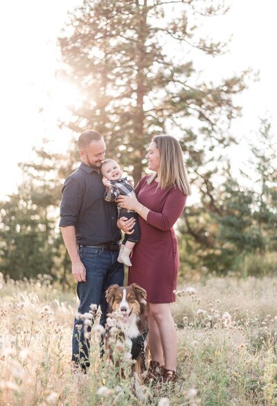 family outdoor photos with parents sitting in the mountains with their toddler son and medium sized dog smiling while in a field together captured by denver family photographers