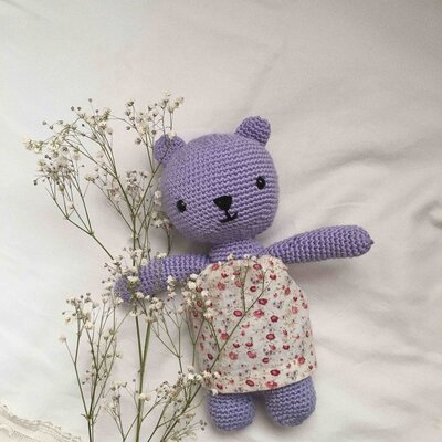 A knitted purple teddy bear is framed in the center of the image, against a white sheet in the background. Two sprigs of baby's breath flowers are also visible next to the bear.