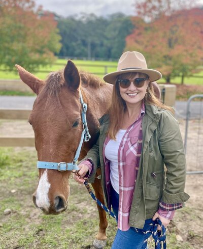 A women with a brown hat on, brown top and blue pants, sitting on a dark brown horse smiling. 