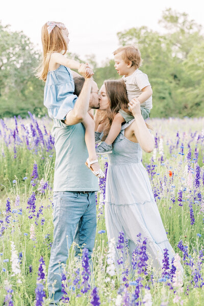 family of 4 in wildflower field in richardson with mom and dad kissing and girl and boy on their shoulders for family photos