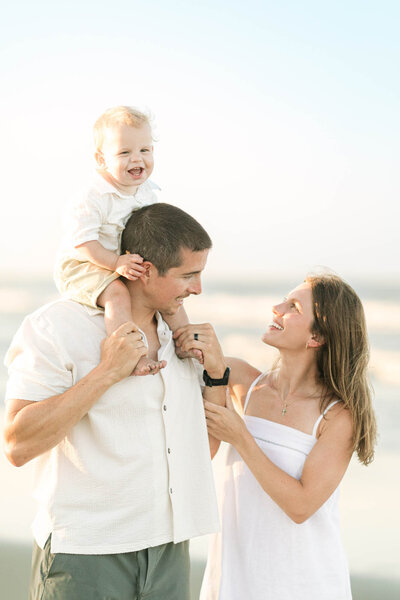 A family photo on the beach of mom and dad looking at each other and their son sitting at dad's shoulders and laughing