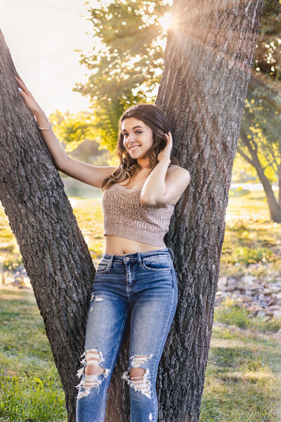 A senior girl posing in front of a tree. Senior is holding her hair in one hand and placed her other hand on the tree.