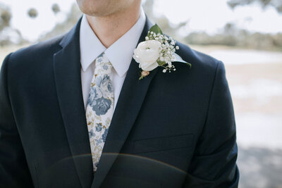 Close up of groom's navy suit and floral white and navy long tie with ranunculus boutonniere pinned on lapel.