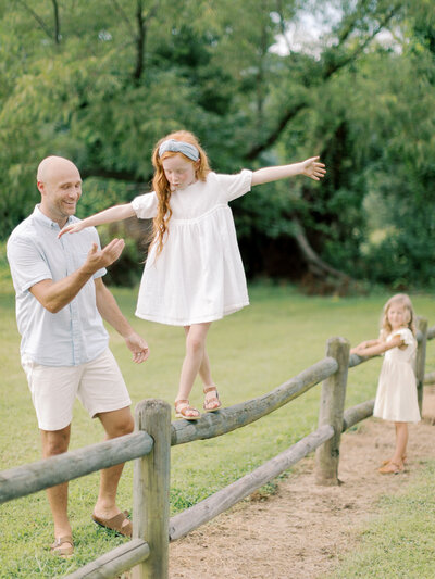 Father smiles and offers support to young red-headed daughter in white dress walking atop a wooden fence in a green field while younger sister watches taken by Little Rock photographer Bailey Feeler Photography