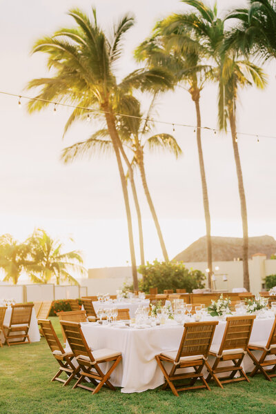 An outdoor wedding reception setup with round tables draped in white linens, surrounded by wooden chairs. The venue is set on a lawn with palm trees and string lights, as the sun sets in the background