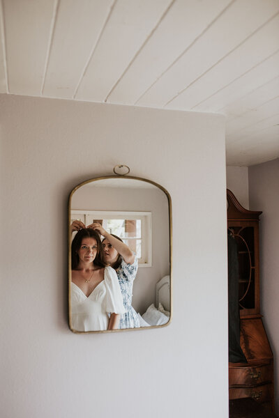 a bride looking in the mirror having her maid of honor put her veil in