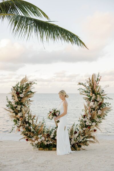 Bride standing in front of a floral arch on the beach, captured by Claudia Amalia Photography in Miami, Florida.