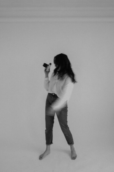 blurred black and white photo of female, Aislynn Palmer Wedding photographer, with long brown hair, holding a camera wearing a white button-up with her face hidden in a white studio in Winnipeg MB.