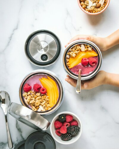 Hands preparing raspberry and mango smoothie bowls topped with granola in stainless steel containers, representing nutrient-dense, hormone-supportive meals.