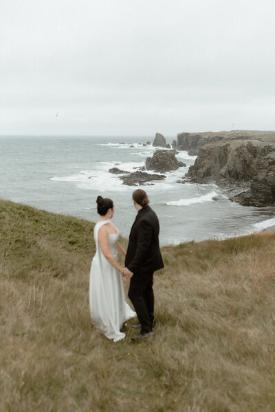 bride and groom hold hands on a grassy hill and look out at the sea stacks behind them in Bonavista