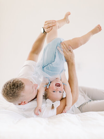Blonde dad in white shirt lays down on white bed while holding giggling one year old daughter in blue dress upside down at Little Rock studio taken by Bailey Feeler Photography