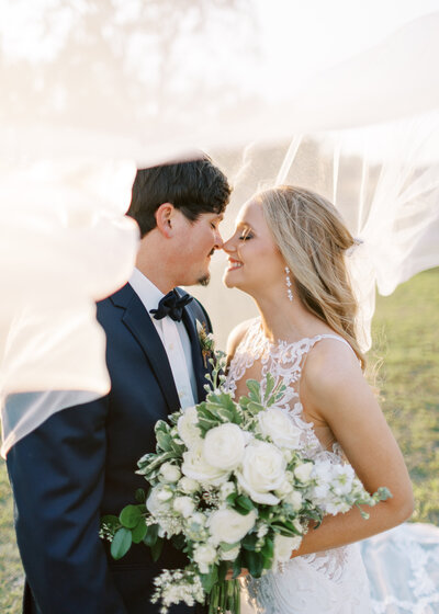 Bride in lace high neck gown and groom in navy tux with bowtie touch noses under a wind tossed veil. An array of white florals are tied in with lush greenery bouquet in the foreground of the image.