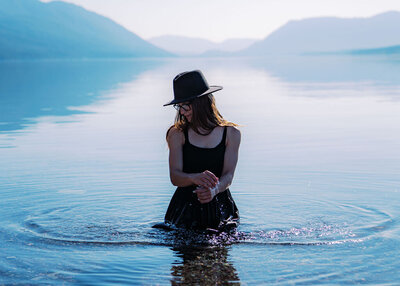 Woman kneeling waist-deep in a calm Lake McDonald wearing a black dress and hat, captured by Joseph Mayden during a creative and adventurous outdoor portrait session.