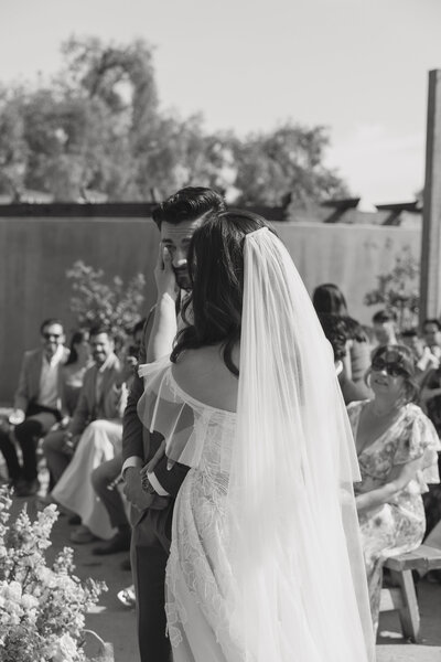 A joyful bride and groom walk along a scenic wildflower trail in Laguna Beach at Top Of The World  during their romantic spring garden wedding. The bride’s flowing lace gown and colorful bouquet complement the lush yellow blooms and sweeping valley views, creating an intimate and picturesque outdoor celebration.