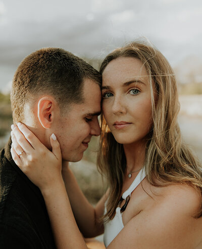 Close up photo of a couple, one looks at the other, one looks at the camera.  The person on the right is wearing a white top and has medium brown hair. They look to be on a beach, the sky is overcast. 