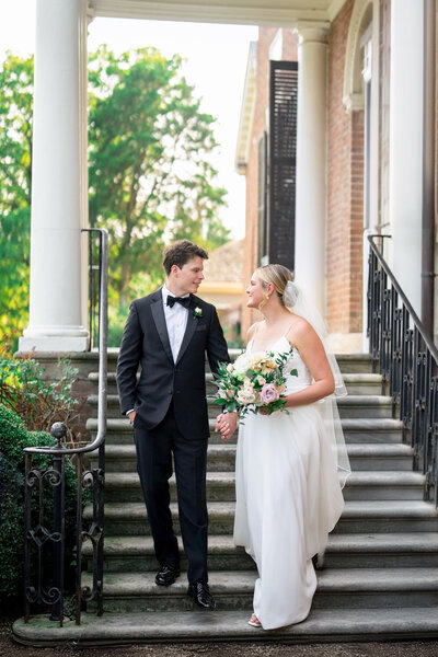 A couple lovingly gazes at each other and walks down steps in a garden setting with a brick building in the background. The bride wears a white gown and holds a bouquet, and the groom wears a black tuxedo.