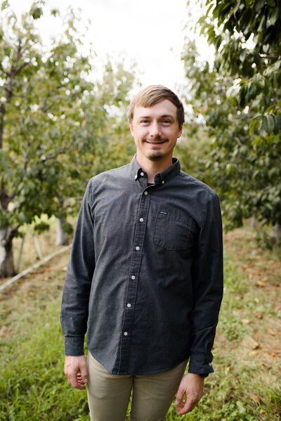 A smiling young man standing in the middle of two apple orchard rows. He's wearing a dark blue button-up shirt and khaki pants