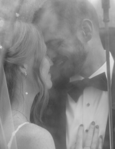 A bride and groom share a kiss in front of a window, dressed elegantly for their black tie wedding.