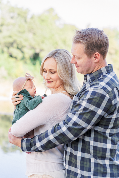 A couple stands in front of a river at Blue Hole Park. The mother holds their newborn baby. The father stands behind the mother and wraps his arms around her. They both look at their baby with a soft smile on their face in this tender moment captured by a photographer specializing in newborn photography in Austin, TX