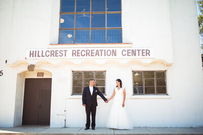 Bride and Groom hold hands and look at one another in front of the Recreation Center at Hillcrest Park