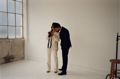 Couple in pantsuits stand close together while looking through photos on a camera