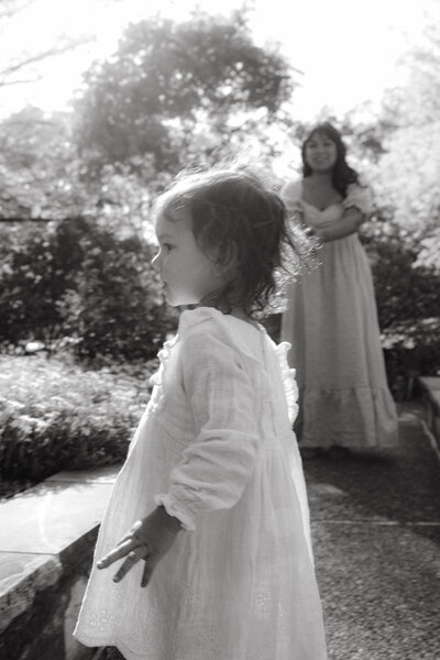 Close-up of a daughter looking at flowers in a garden with her mother blurred in the background, captured in a documentary-style family portrait by Emily Ambie.
