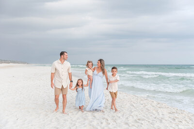 Young family walking along the beach together, dressed in coordinated light blue and neutral outfits, parents holding children’s hands with waves in the background.