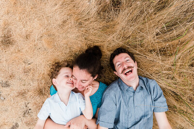 Family laying in brown tall grass next to each other, mom holding son on top of her tickling him, all with smiles and lauging