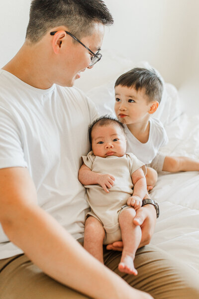 Portland Newborn Photographer_ A photo of a dad holding his newborn baby and smiling at his toddler son. In home lifestyle newborn photos 