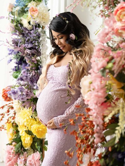 A pregnant woman in a lavender lace dress stands amidst vibrant, cascading flowers in pink, yellow, and purple tones, exuding a serene and joyful mood.