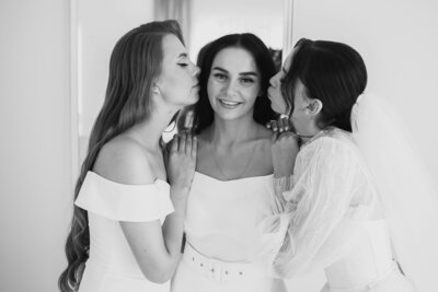 Smiling bride in white dress with two bridesmaids kissing her cheeks in a soft black and white wedding photo