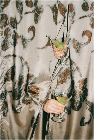 Two hands stick out from behind a brown curtain holding martinis