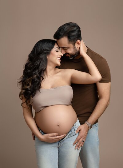 Pregnant Mom in jeans and a tube top holding on to dad and smiling and holding her baby bump. Photo taken at Atlanta Maternity photo studio in Woodstock GA.