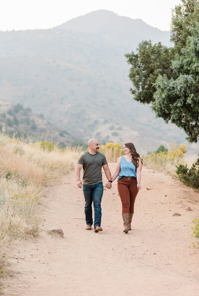 denver couples photography with man and woman holdin ghands togegther for their outdoor engagement session in the mountains as they walk together on a large dirt path with the rockies int he distance