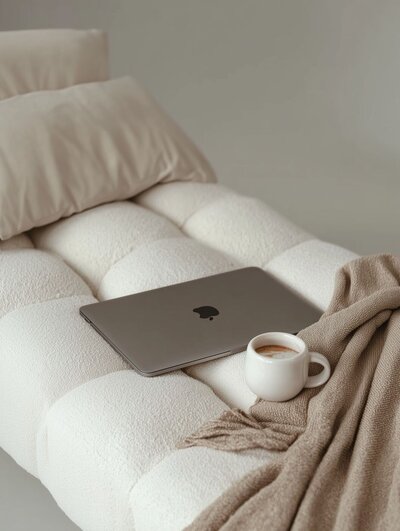 Cozy neutral lounge chair with a folded beige throw, closed laptop and a mug of coffee resting on the cushioned seat