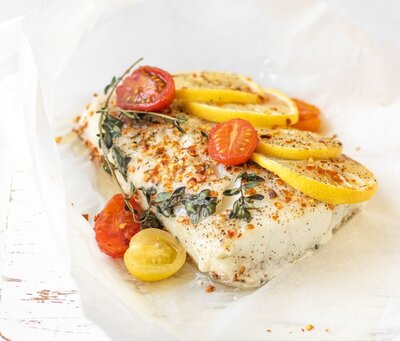 A close-up of fertility-friendly Mediterranean cod baked in parchment, topped with slices of fresh lemon, halved cherry tomatoes in red and yellow, and sprigs of thyme for a nutrient-rich, antioxidant-packed meal. The fish is seasoned with herbs and spices, resting on white parchment paper with a rustic white wooden background.