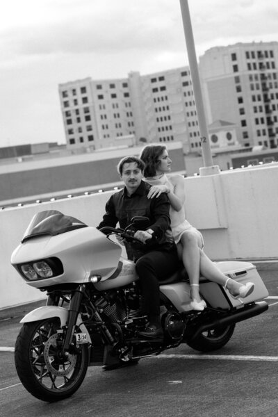 A couple looks at the camera in a black & white photo while sitting on a motorcycle