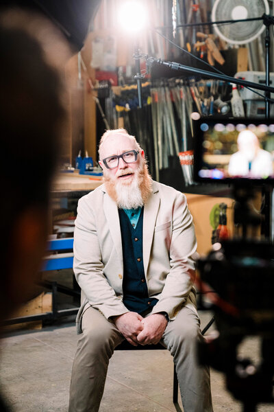 a man with a red and grey beard sits under a boom microphone in front of a camera