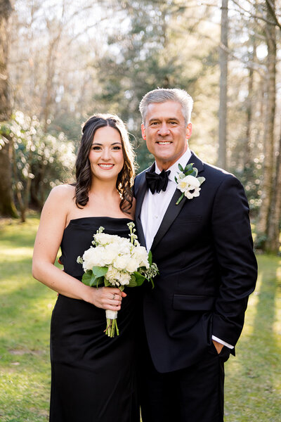dark haired girl in black dress holding flowers standing next to grey haired man in a tux