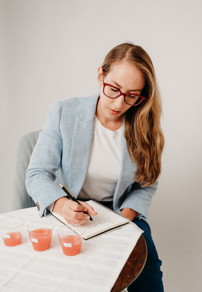 Rachel Toner wearing a blue blazer and white T-shirt and taking notes in a notebook next to pink drinks