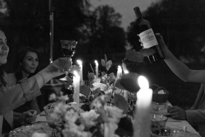 lack and white image of hands raising wine glasses in a toast at a romantic outdoor dinner table with candles and floral arrangements.