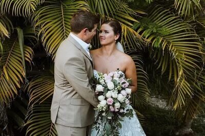 A bride and groom gaze into each other’s eyes in a lush tropical garden, with the bride holding a bouquet of white and blush flowers at their destination wedding.