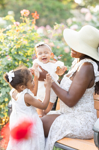 A mother holding a young toddler girl in a sun light rose garden as the toddler admires a soft pink rose she holds in her hand. 