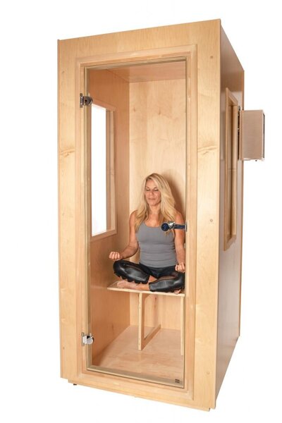 A small, wooden salt therapy booth with a glass door and side window. Inside, a simple wooden chair and a small bench offer a quiet retreat, reminiscent of a cozy massage therapy space in Tolleson, Arizona. Set against a plain, light background.