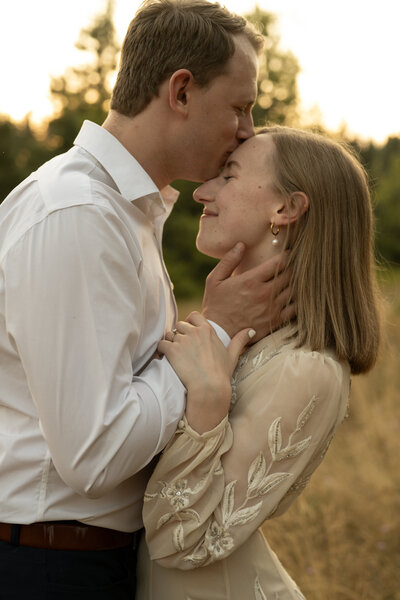 engaged couple in a field with man kissing his fiance's forehead