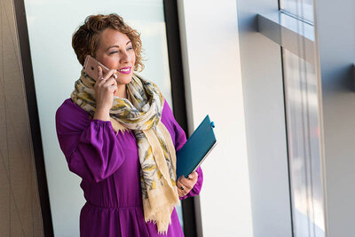 This image depicts a woman engaged in a phone conversation. She has curly short hair and is wearing a purple dress with a gathered waist, complemented by a patterned scarf in shades of yellow and beige. Her expression is cheerful, suggesting a pleasant interaction. In her left hand, she holds a closed teal notebook, indicating she may be in a professional setting or conducting business. The background is light and features modern architecture, possibly the interior of an office building. The woman's attire and accessories suggest a sense of personal style and professionalism.