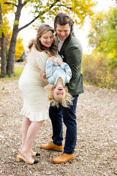 A mom and dad hold their toddler daughter between them and tip her upside down as she laughs and they smile at her.